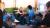 Participants sit in a circle in a lounge area of the pub venue, taking part in one of the interactive activities. One of the participants is seated with Ash, a puppet figure, on a sofa, shake hands while others observe and engage in the group exercise. Lamps, chairs and green wall panelling are visible in the background.