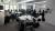 Conference participants sit around several tables arranged in a workshop-style layout in a bright meeting room. One participant at the front of the room addresses the group while attendees listen and take part in the discussion. Water bottles, notebooks, and personal belongings are placed on the tables. Large frosted windows provide natural light, and the room features white walls, grey carpeting, and presentation equipment including a flip chart and display screen.