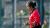 Ruchitha Venkatesh in a red and black Hong Kong, China team uniform stands on an outdoor cricket field, watching a cricket ball suspended in mid-air directly in front of them. The player is shown in profile, with hands positioned beneath the ball as if preparing to catch or control it. The background is softly blurred, highlighting the player and the ball, with fencing and greenery visible around the sports ground.