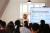 A speaker giving a presentation in front of a large screen, with the backs of students in the foreground.