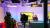 A woman sits at a news desk in a brightly lit studio. A camera operator stands nearby. A screen displays "University of Westminster." The mood is professional.