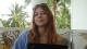 Alexandra Bukhareva sitting in front of a laptop smiling. Behind her there are green palm trees.