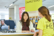 A student stands at a university information desk speaking with a campus tour guide wearing a yellow T-shirt. A sign behind them reads “Campus Tours,” and other people and information stands are visible in the background of a busy indoor student fair. If you want it