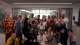 A group of about 25 people poses indoors for a photo, smiling and raising their hands, with a Pride flag hanging in the background.