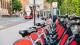 A row of Santander bikes parked on a pavement with a bus in the background.