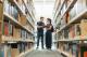 Two students standing between library shelves, looking at a book together in a bright academic library.