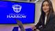 Alumna Kareena Ravaliya posing for a photo in front of her design of the Harrow Logo on a screen behind where she is sitting.