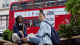 Two students sit outdoors on steps with backpacks, with a red London bus passing behind them.
