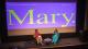 Gemma Cairney (left) and Laura Bates (right) sit on chairs on a stage in front of purple cinema screen with Mary in large green text.