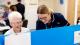 An elderly woman and a nurse are looking at something together, which is covered by a blue corkboard.
