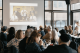 People seated at tables watching a video projected on a large screen in a bright room.
