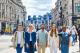 Five diverse young adults smiling and walking together down a busy London street, with historic buildings, Union Jack–style event banners, and an Underground sign visible in the background.