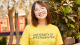 A smiling student wearing a yellow “University of Westminster” T-shirt stands indoors among plants.