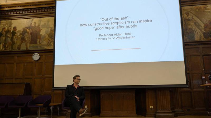 Aidan Hehir delivering his inaugural lecture in front of a projector screen reading the lecture title: “Out of the Ash”: How Constructive Scepticism Can Inspire “Good Hope” After Hubris