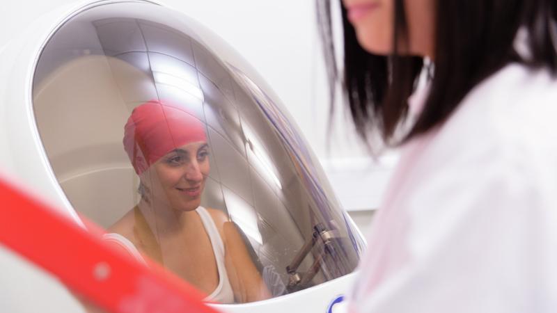 A female athlete sitting in a pod with a glass window, whilst a female in a white coat attends outside the pod