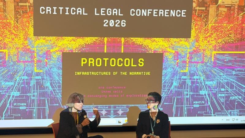 Ingrid Guardiola Sánchez and Julia Chryssostalis seated on stage take part in a discussion in front of a large projected conference slide. The screen displays the text “Critical Legal Conference 2026” and “Protocols: Infrastructures of the Normative,” with a colourful digital graphic background. Both speakers hold microphones while seated facing each other, and the presentation fills the backdrop of the conference venue.