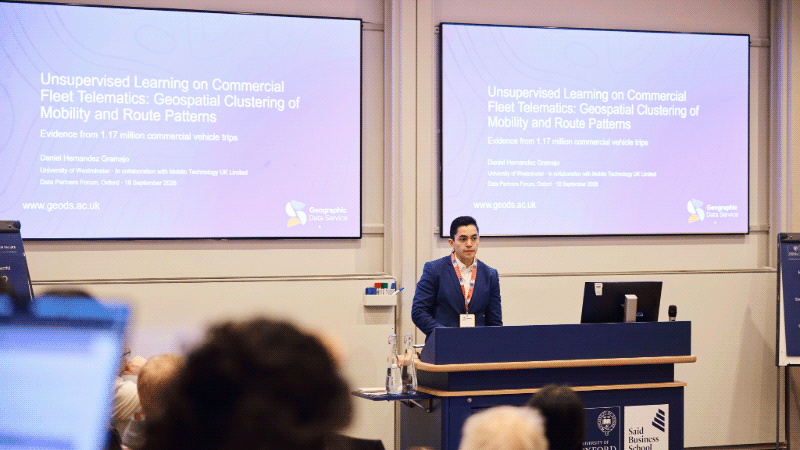 Westminster Master's student presents his research on geospatial clustering of mobility and route patterns at the Data Partners Forum in Oxford, standing in front of two presentation screens.