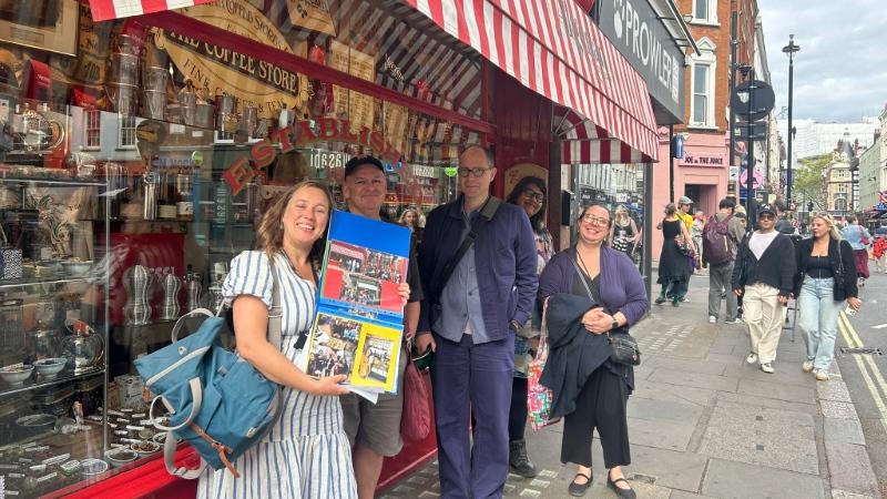 Dr Emily Falconer stands with walk participants outside a shop on a busy London street, holding a display board featuring photographs