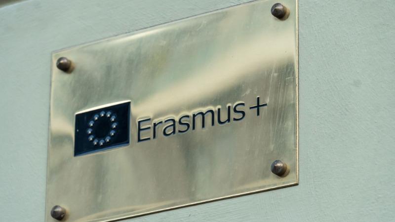 Close-up of a metallic plaque mounted on a light-coloured wall displaying the Erasmus+ logo and name. The rectangular plaque is secured with four visible screws at the corners and features the Erasmus+ text beside the European Union emblem.