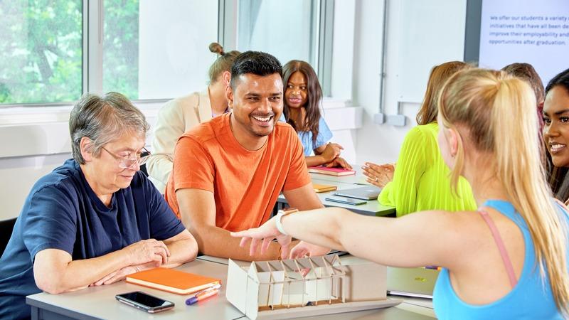 Participants sit around a table in a bright classroom, discussing a scale architectural model during a collaborative workshop. Notebooks, pens, and laptops are visible on the tables, while large windows provide natural light to the room.
