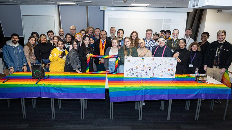A large group of students and academics behind a table with a bright rainbow tablecloth