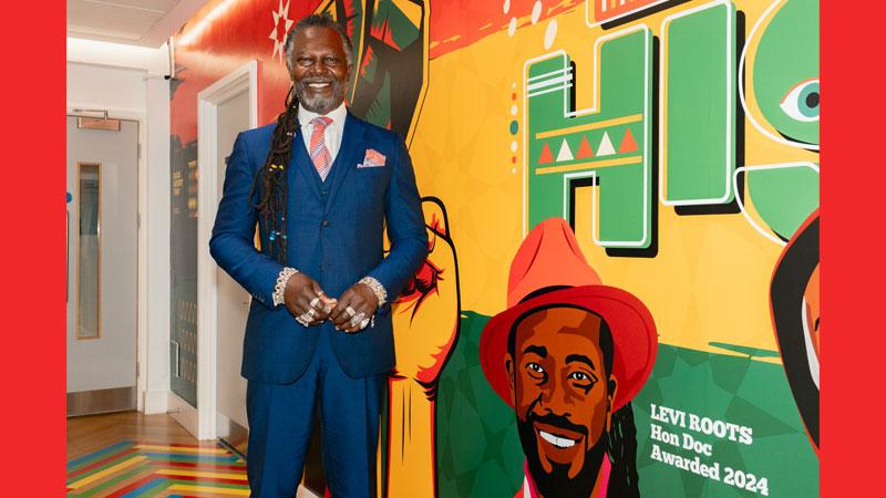 Levi Roots stands smiling next to his portrait on the Black History wall mural.