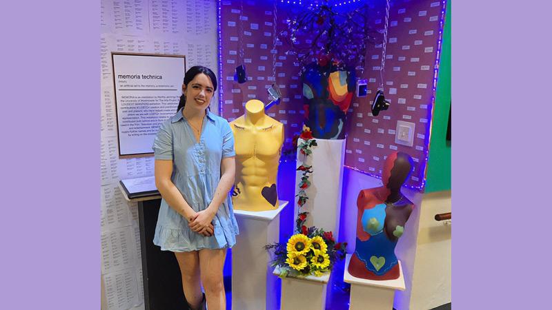 Martha Jennings next to her creation Memoria Technica in corner, featuring three torsos of different genders on plinths, decorated with a variety of flowers and blue led lights, on the wall name stickers of LGBT+ people from art industries