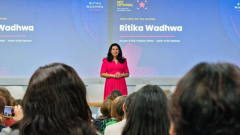 Ritika Wadhwa stands at the front of a lecture theatre addressing an audience seated in rows. Behind the speaker, large presentation screens display a slide featuring the text “Ritika Wadhwa” and information about the author and business leader. The speaker wears a bright pink outfit and stands centre stage, while audience members are visible in the foreground facing the presentation. The room is brightly lit, with the presentation projected across multiple screens at the front of the venue.
