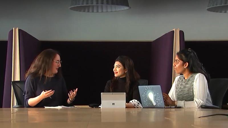 Three students sat at large desk talking to each other with laptops