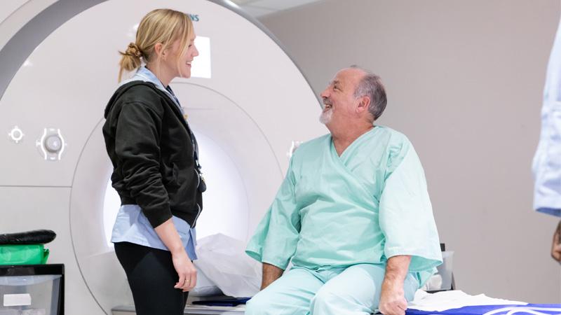 Picture of a man in green hospital robes preparing for an MRI scan with a nurse.