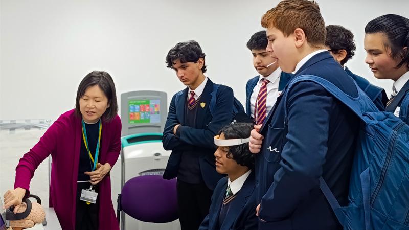 A group of school pupils are being shown an experiment on the brain. One pupils is wearing a brain scanning device and the other pupils are watching behind him in amazement.