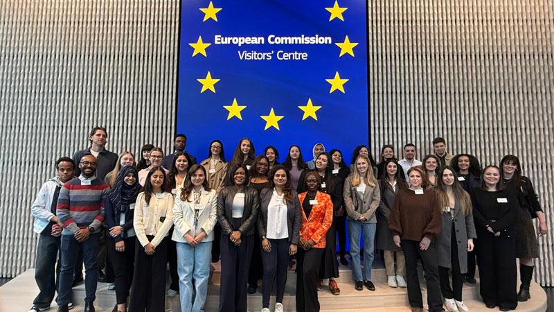 Group photo of student attendees at the European Commission visitor centre. They are standing in front of a blue EU flag.