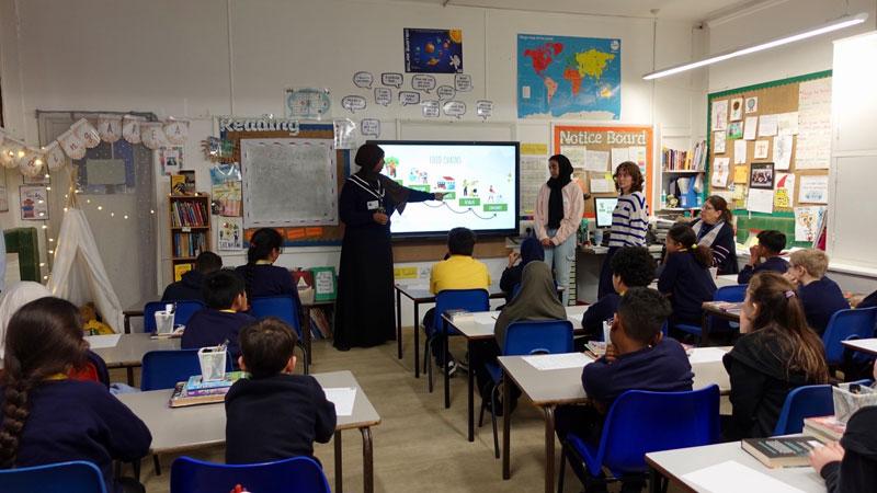 Westminster students delivering a workshop to pupils sat in rows of desks at Queens Park Primary School.