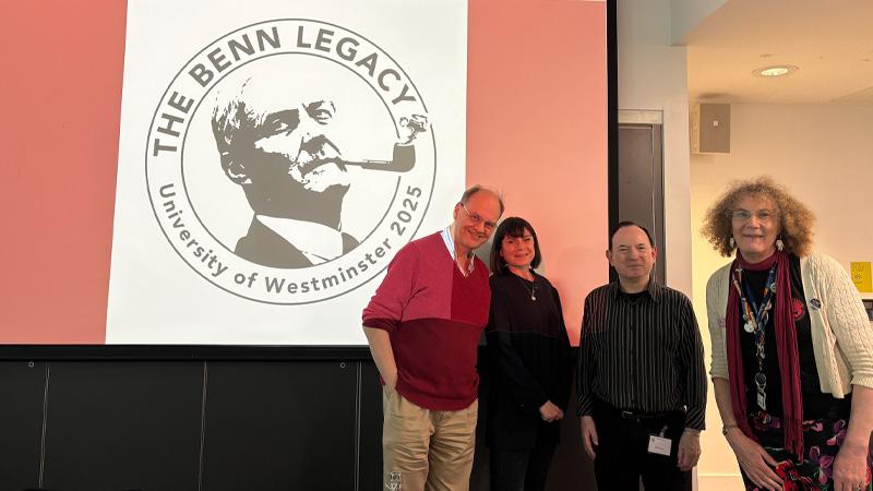 Photo of the organising team of The Benn Legacy event standing in front of a projector displaying the conference's logo, a black and white picture of Tony Benn smoking a pipe surround by the text THE BENN LEGACY UNIVERSITY OF WESTMINSTER 2025