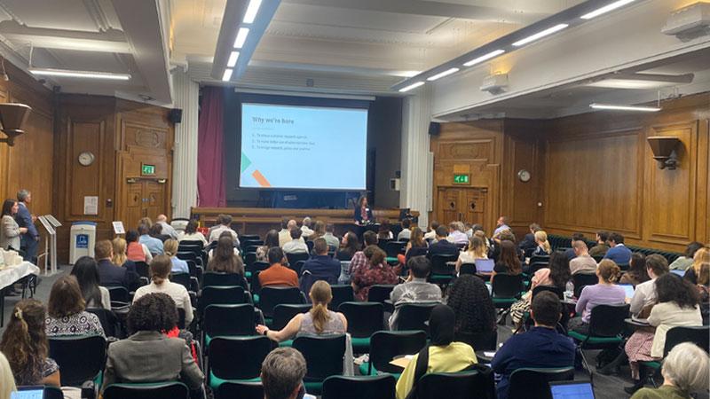 Photo of Karen Powell from ADR introducing the conference to a full audience. Behind her is a projector screen which reads: Why we are here and various reasons why the research is important.