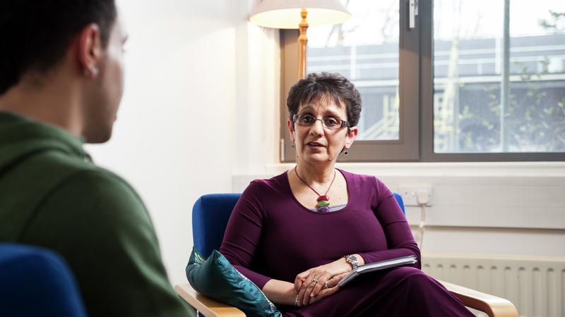 A male and a female sat on chairs facing towards each other in a room, the female has a clipboard and is listening to the male.