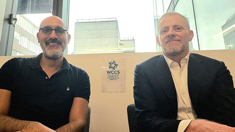 Dr Nikolaos Souvlakis and Professor Damien Ridge taking a photo in front of the Westminster Community Counselling Service logo