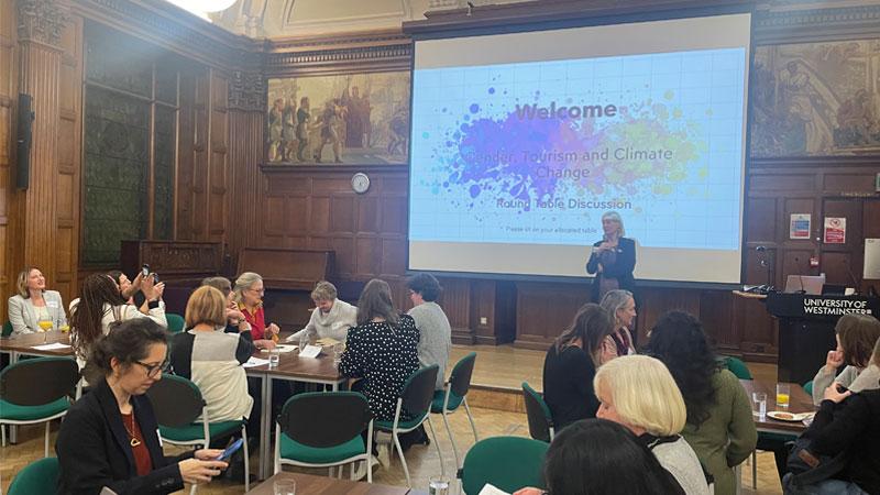 Attendees at the Gender, Tourism and Climate Change sit around tables in front of a projector with "Welcome" "Gender, Tourism and Climate Change Round Table Discussion" displayed on it. Dr Stroma Cole stands at the front.