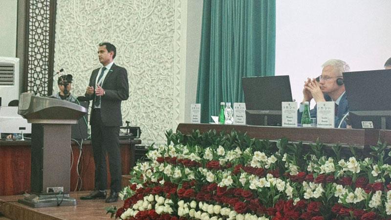 Mohammed Gulrez Zariwala delivering the welcoming speech at the conference. He is standing at a podium on stage where the panel are sat next to him on a table surrounded by flowers.