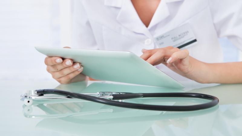 A woman wearing white, using an ipad, with a stethoscope on the table
