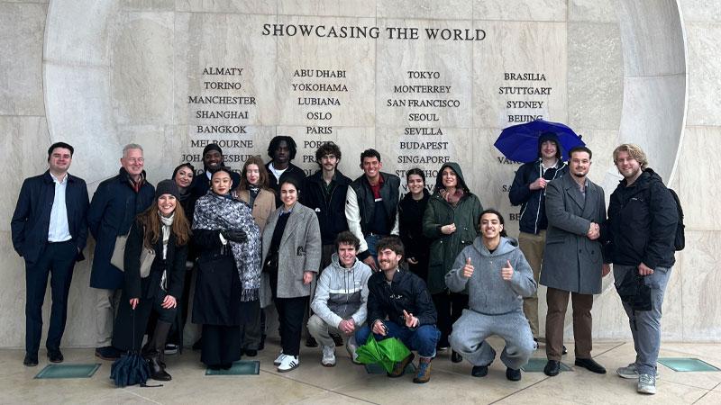 Group photo of students on trip to JCDecaux headquarters in front of a wall that reads SHOWCASING THE WORLD with lots of different countries