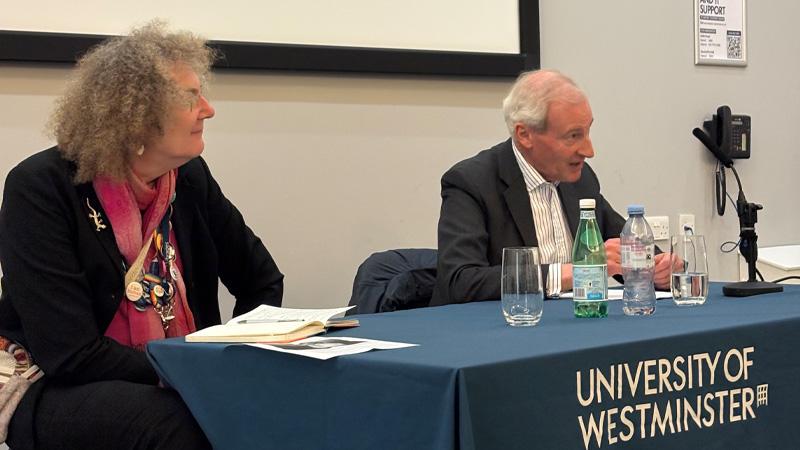 Professor Catterall and Sir Laurie Magnus sat at a table with a tablecloth that says UNIVERSITY OF WESTMINSTER