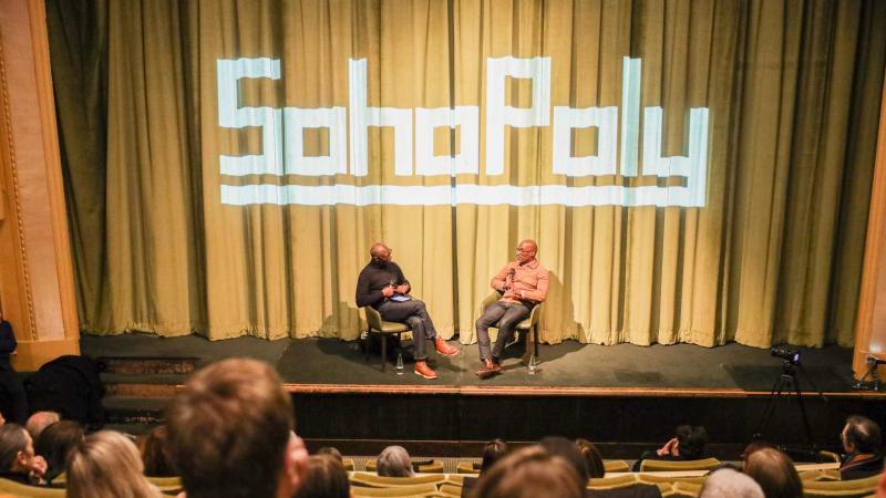 Dr Mykaell Riley leading the Q&A session with Clint Dyer on the stage of the Regent Street Cinema with the Soho Poly logo projected onto the curtain behind them