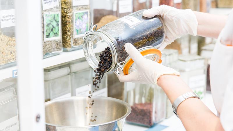 Hands pouring a glass jar full of herbs into a bowl, with a row of jars full of herbs in the background