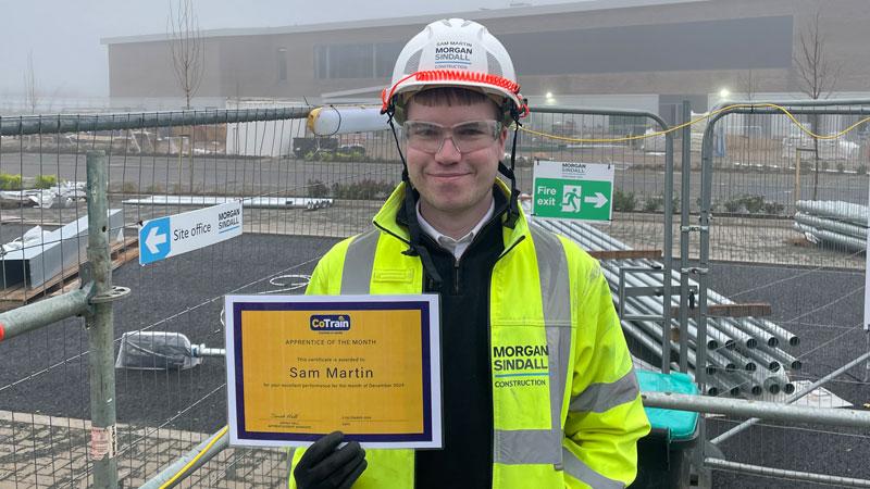 Sam Martin on a building site wearing a hard hat and high-vis jacket and holding his Apprentice of the Month certificate.
