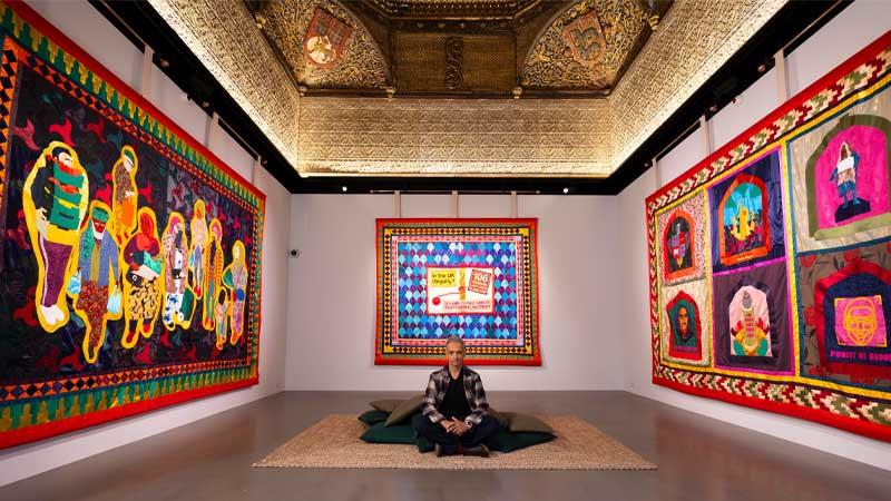 Shaheed Saleem sitting on the floor in the middle of his installation, surrounded by tapestries and under the Torrijos ceiling