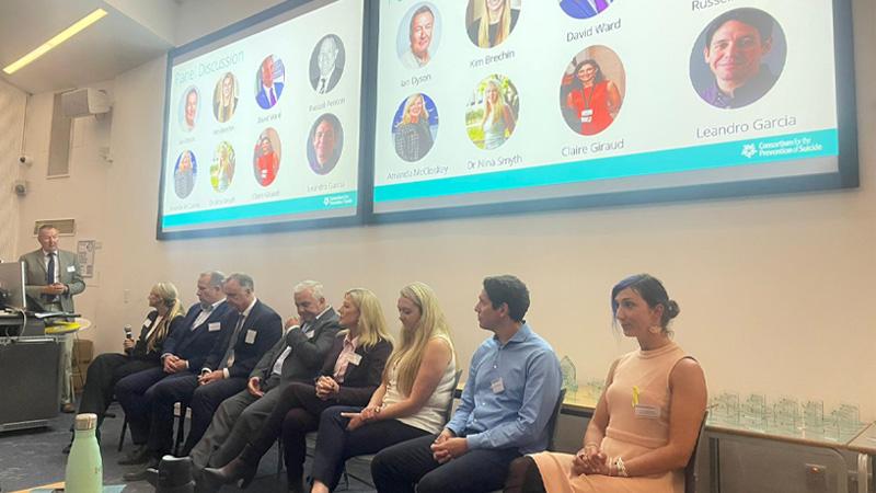 Photo of the panel at the Suicide Prevention Day conference. Behind them are screens showing headshots of the panellists.