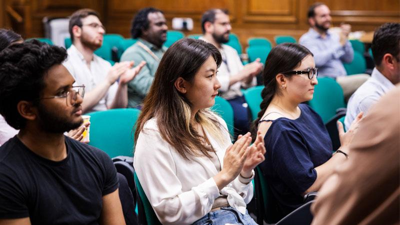 Picture of the audience clapping. They are sat on green chairs in a wood-panelled room.