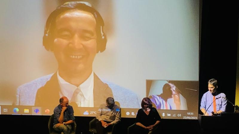 Picture of speakers sat on stage at the Transition Economies Event with the Vice-Chancellor speaking to the right of them at a lecturn and the face of a speaker projected on the screen behind
