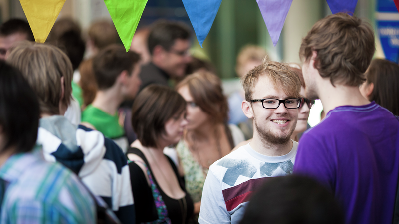 male student smiling at camera, surrounded by other students, and colourful bunting above him.